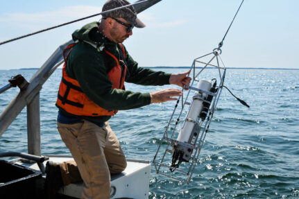 Research kneels on a boat setting up a measuring device to be placed in the water.