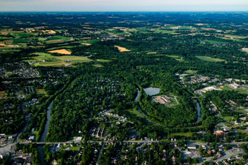 An aerial view of a river flowing through green trees and houses. In the far left, you can see farm fields