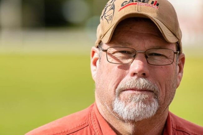 Farmer with glasses, red shirt, and a hat looks over his property.