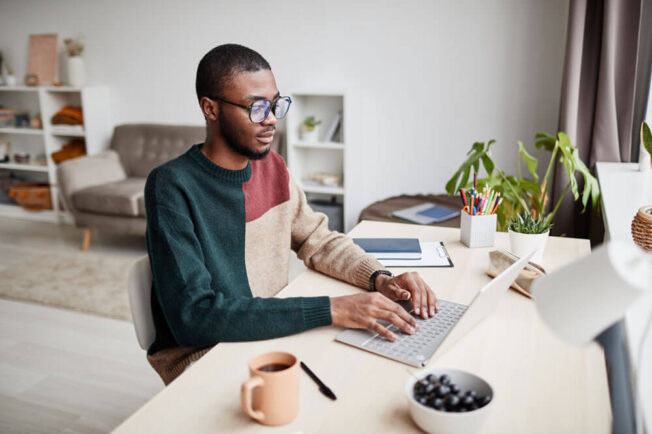 Young black male sits on a desk at home working on his laptop.