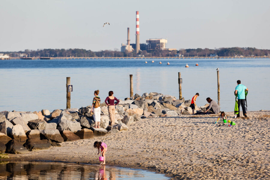 Smokestacks are seen from across a river, where people play along a beach in the foreground.