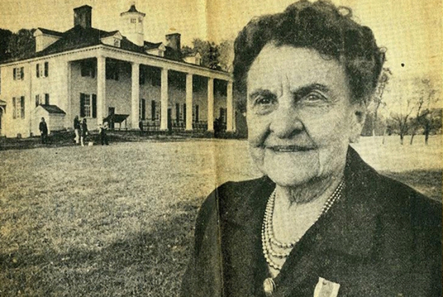 Women with dark blazer on stands in front of an old white home with white pillars and a black roof.