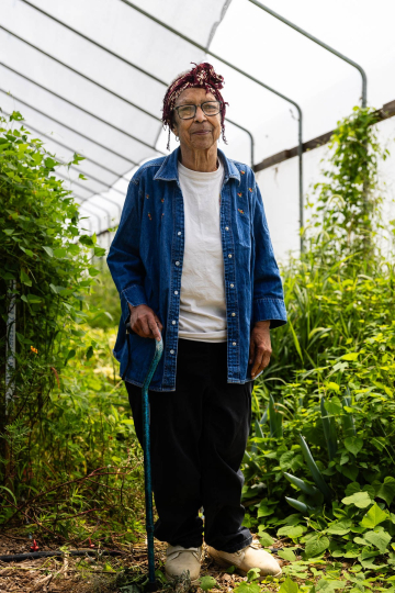 Ira Wallace is an elderly Black woman wearing a jean jacket and red bandana, posing for a portrait in a greenhouse filled with lush, green growth.