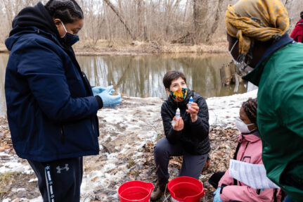 Three volunteers stand near the side of a river and review water quality samples.