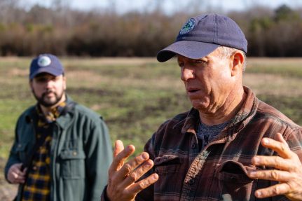 Two men, wearing the same blue baseball hat, standing outside in a farm field. The older man speaks, gesturing with his hands. To his left, the younger man listens.