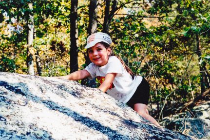 A young child climbs a large boulder. Green trees fill the background.