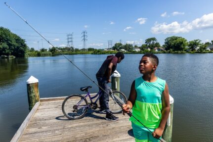 A kid hold a fishing pole and another is on a bike at a fishing pier.