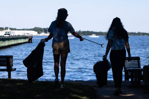 Two women face the Chesapeake Bay, with one holding a trash bag and long trash grabber.