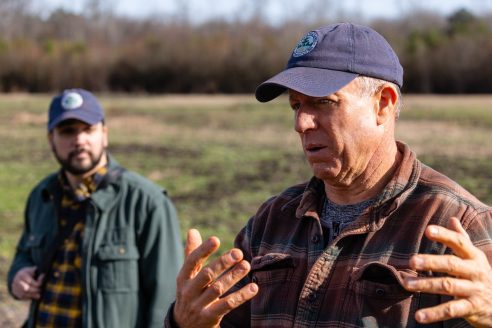 Two men, wearing the same blue baseball hat, standing outside in a farm field. The older man speaks, gesturing with his hands. To his left, the younger man listens.