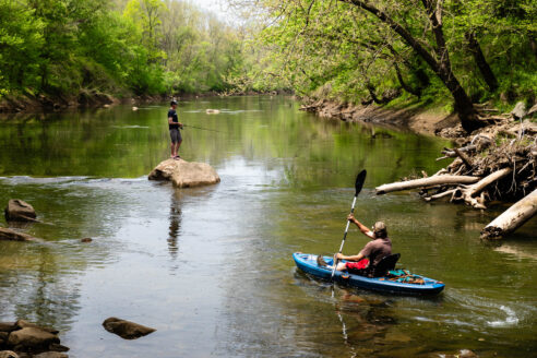 Man kayaks down a creek where someone is fishing.