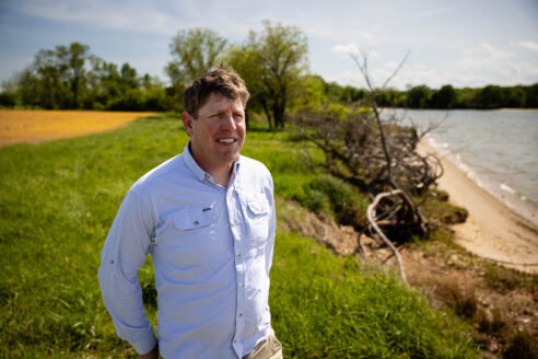 Will Gordon squints in the sun, facing an eroded shoreline with a yellow farm field behind him.