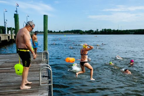 A woman leaps into a river from a wooden dock, joining more than a dozen swimmers who are already in the water.