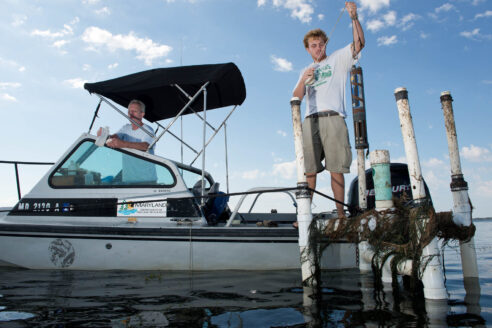 Scientists on a small boat manipulate monitoring equipment rising from the water and covered in underwater plants.