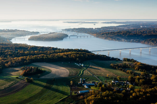 An aerial view of the Susquehanna River flowing toward the Chesapeake Bay, through land dotted with farm fields, forests and bridges.