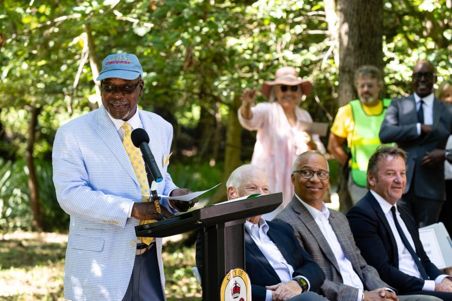 Vince Leggett stands in front of a microphone and podium with event attendees sitting and standing to his left side.  The group is outdoors with trees in the background.