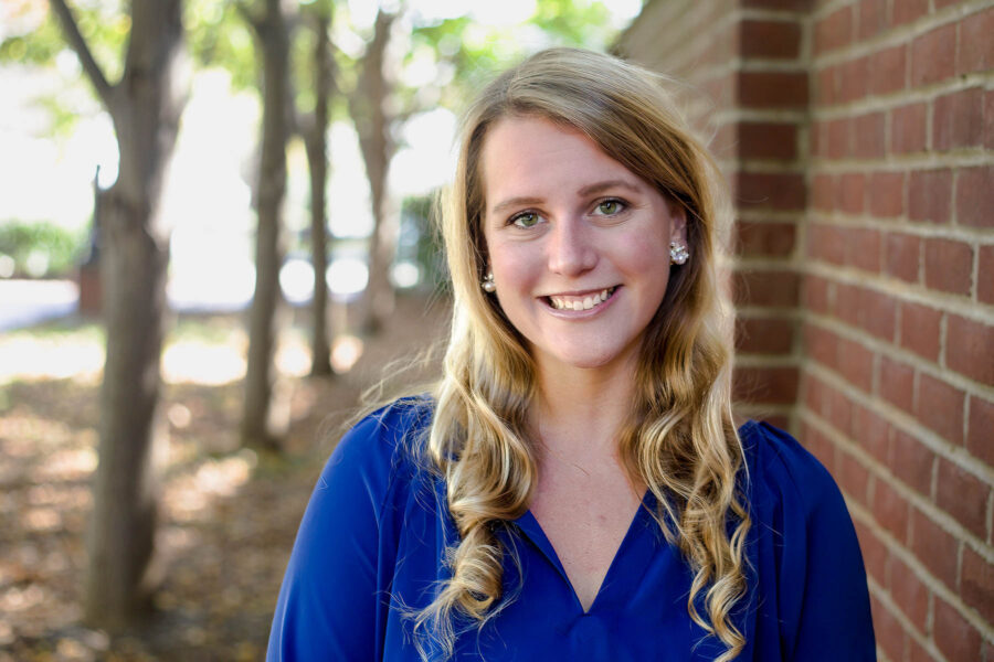 Sara is a white female with blond hair. She is standing outside by a row of trees.
