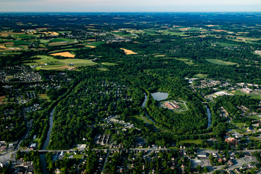 An aerial view of a river flowing through green trees and houses. In the far left, you can see farm fields