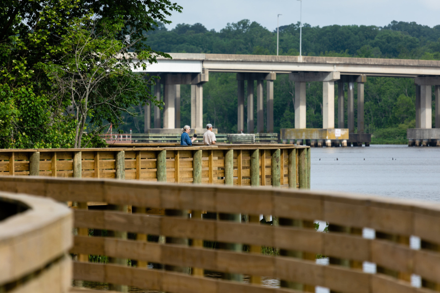 A boardwalk winds over the water with a bridge in the background.