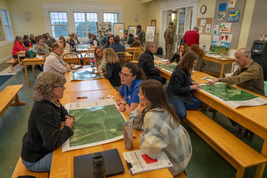 A room is filled with tables and benches. About two dozen people fill the room. People sit on both sides of the table with large maps placed on the tables.