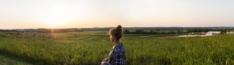 The farmer stands against a wide view of farm fields at sunset.