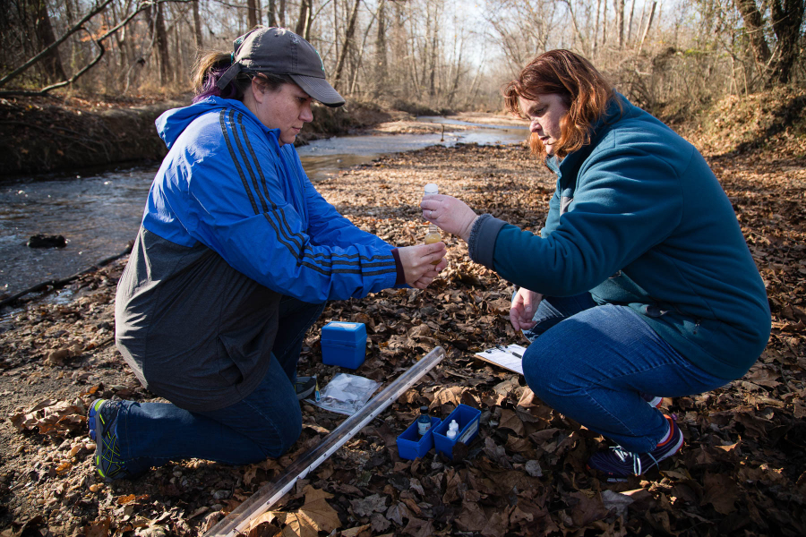 Two biologists kneel next to a stream and prepare water samples.