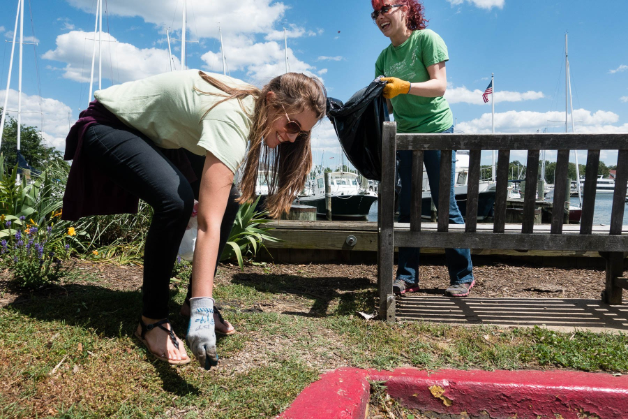 Two women use gloves to pick up trash on a street near boats at a dock.