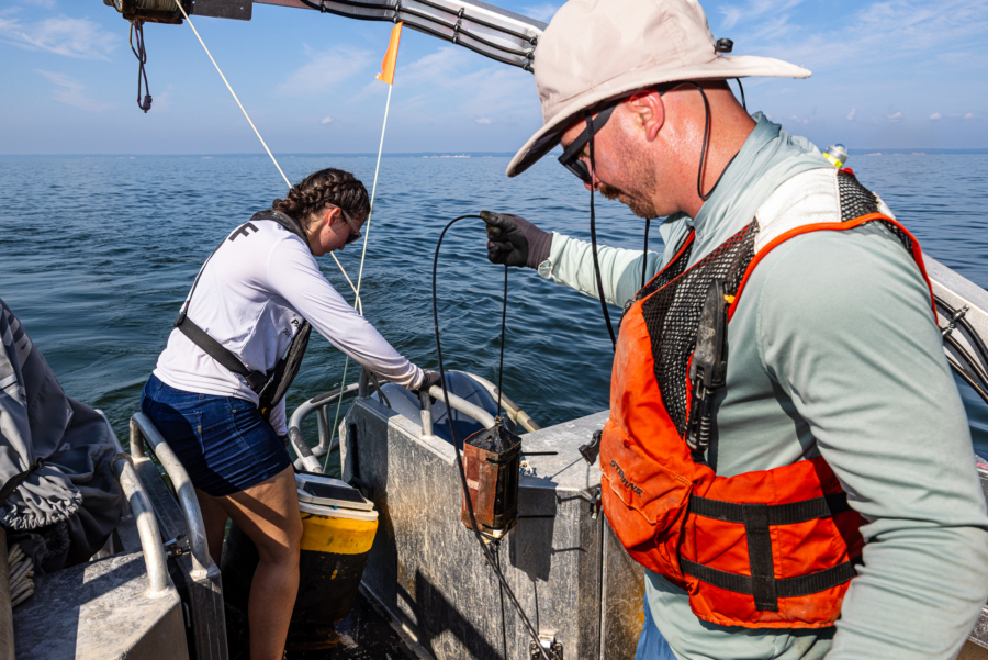 Two technicians hold onto a line as they haul a small yellow buoy onto the back of a boat.