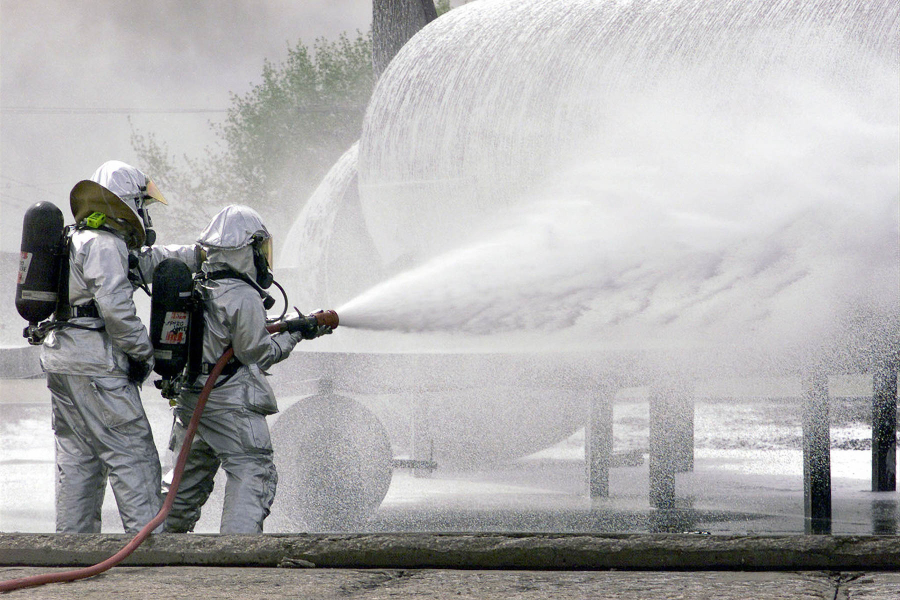 Two men in protective gear spray white foam outside.