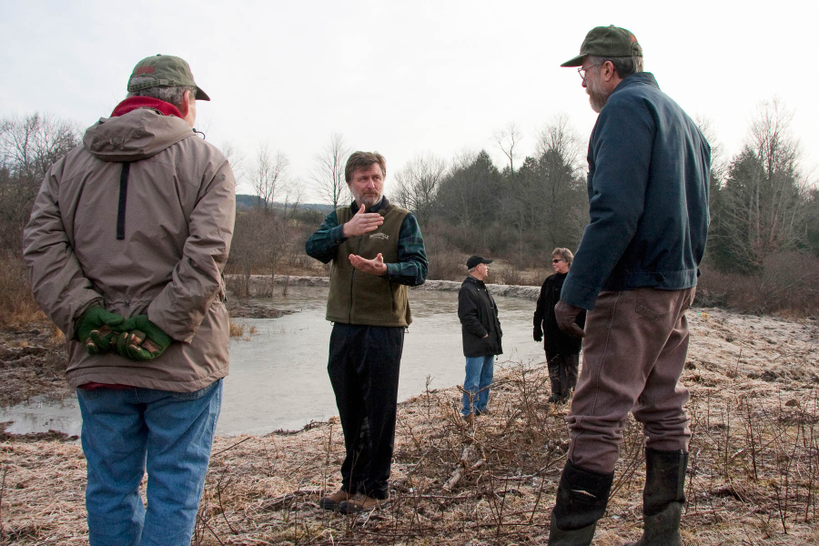 A photo of man gesturing to two others at water's edge on the constructed wetland, with his wife talking to another man some distance away.