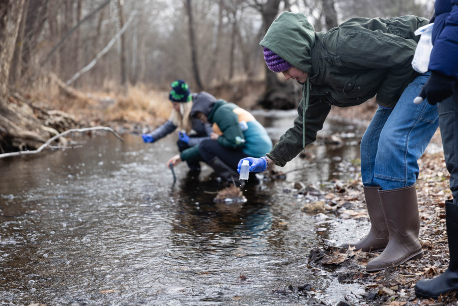 A teenager bundled against the cold holds a water sample above a creek.