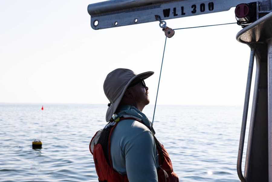 A researcher in a wide-brimmed hat is silhouetted on a small boat with a buoy in the distance.