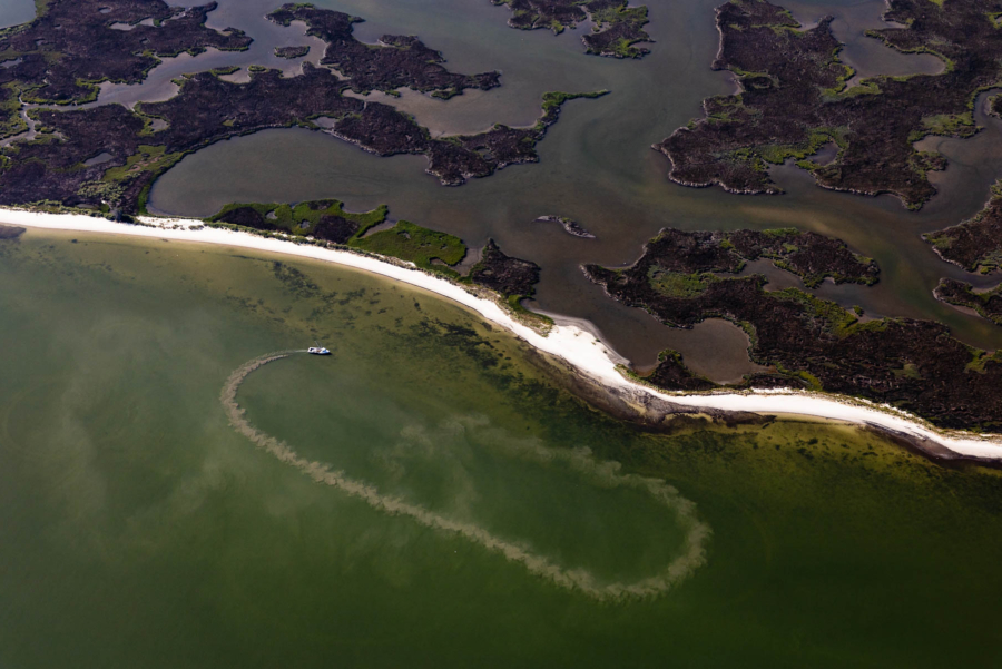 A workboat seen from high above traces an incomplete oval in muddy water next to an expansive marshy shoreline.