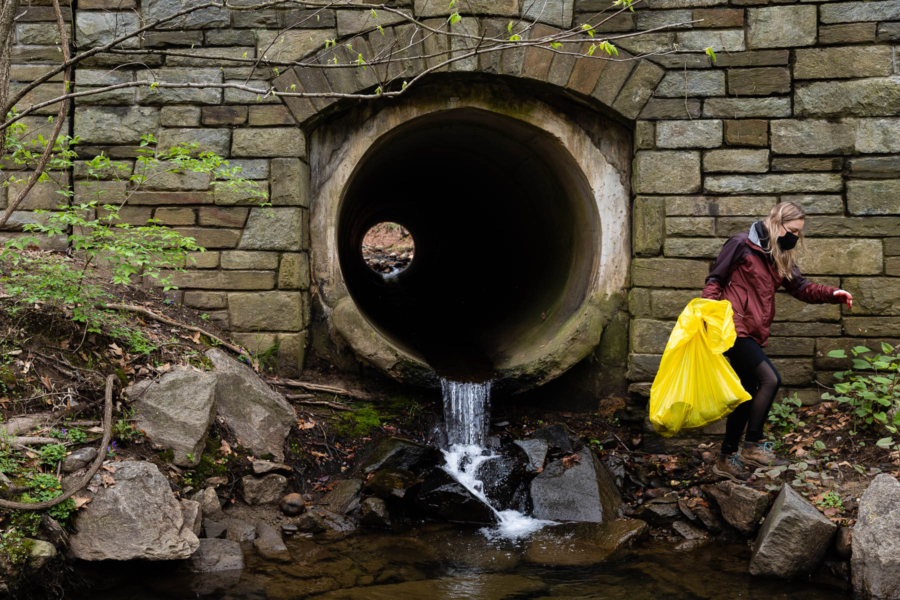 Water flows out of tunnel that is part of a stone bridge. A volunteer holds a bag and picks up trash in front of it.