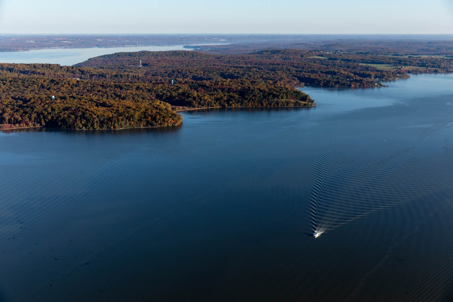 An aerial view of a wide blue river and a forested landscape. In the foreground, a small white boat passes through the river leaving a wake.