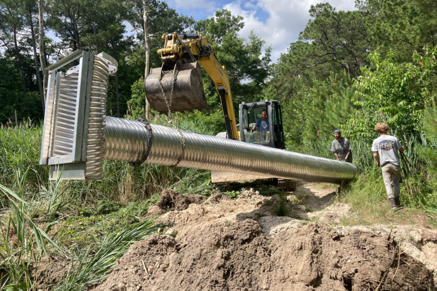 A bulldozer lowers a large piece of metal into a wooded area with green trees. Dirt is in foreground.