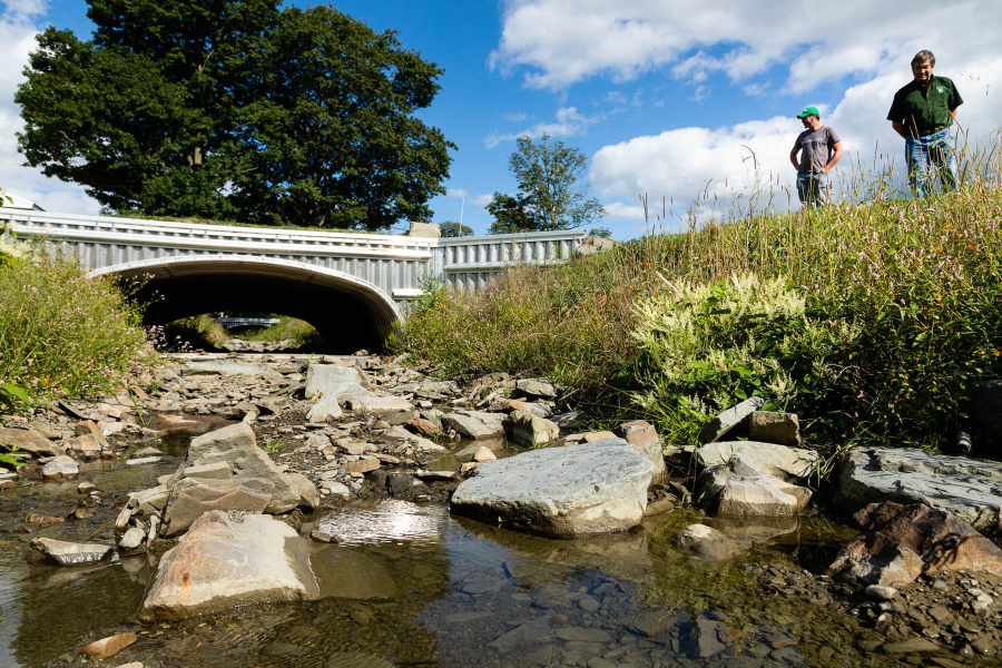 Two men stand on a hillside overlooking a small, rocky stream, which flows uninterrupted through a series of two culverts.