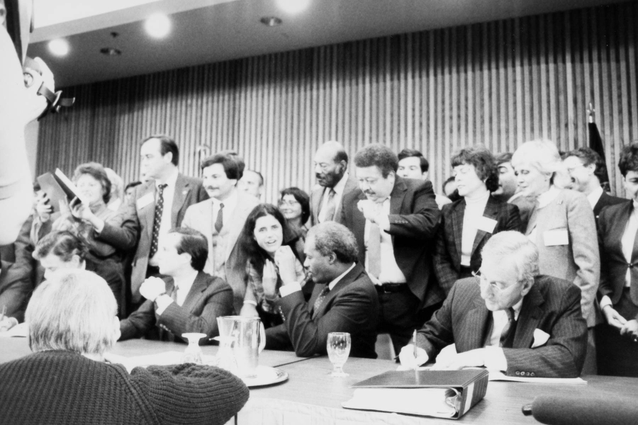 A black and white image of Joseph Gartlan, Jr., then chair of the Chesapeake Bay Commission, sitting at a table while signing the 1983 Chesapeake Bay Agreement amid a chaotic scene of more than a dozen onlookers.