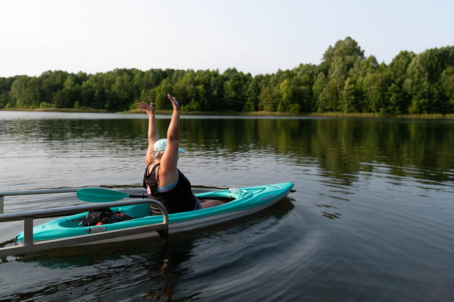 A woman in a kayak throws her hands into the air as she launches her vessel into wide, calm water with forest beyond.