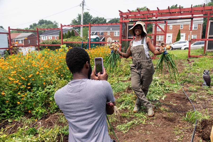 A woman wearing a sunhat and overalls stands in an urban farm's garden plot and proudly holds up two bunches of onions as a man takes her photo with his smartphone.