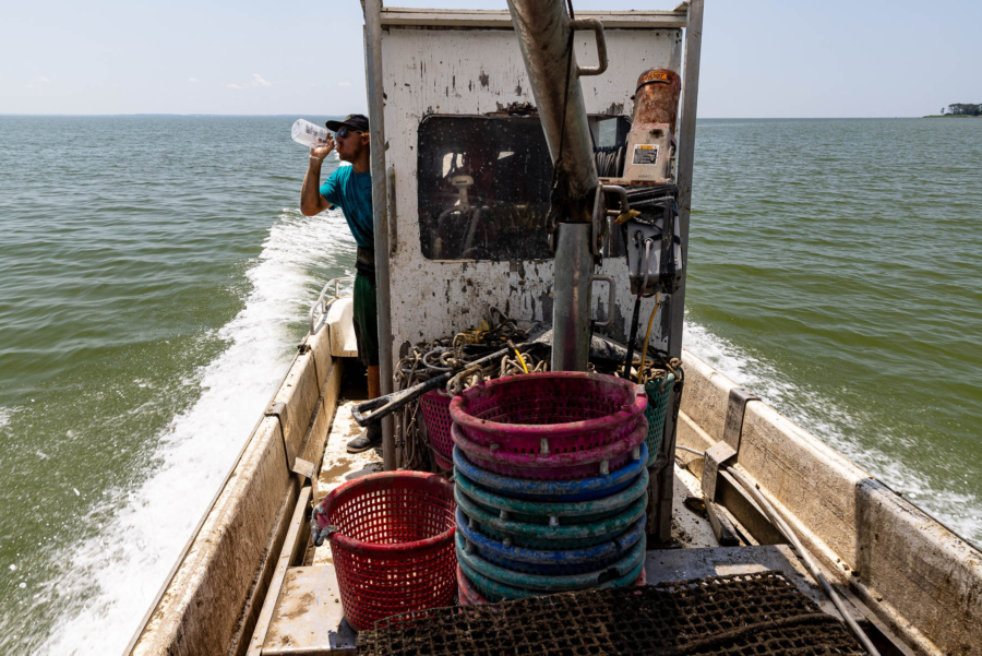 Sturmer tips back a large water bottle under a hot sun, with a wide expanse of water behind his small workboat and a tiny speck of land on the horizon.