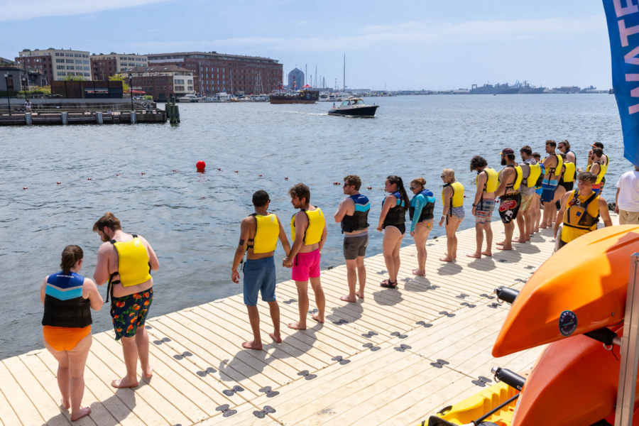 Swimmers line up on the edge of a dock facing Baltimore's Inner Harbor, city buildings surrounding them