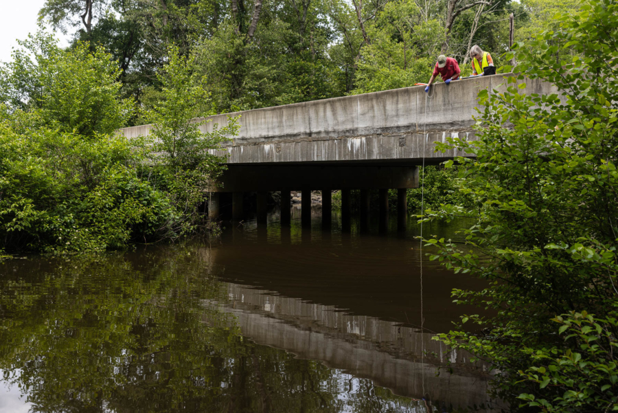 Two volunteer dangle water quality monitoring equipment over a creek to collect samples from a bridge.