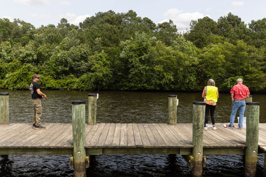 Two volunteers holding equipment share a wooden dock with an angler holding a fishing rod against a forested backdrop on a quiet river.