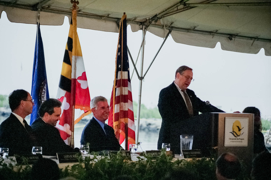 A man stands at a podium with three other politicians sitting down next to him, under a tend on the Bay.