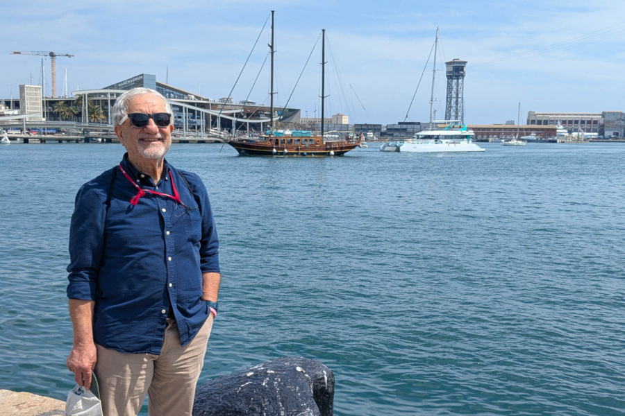 Karimi stands next to a wide harbor with a shoreline and boats in the background.