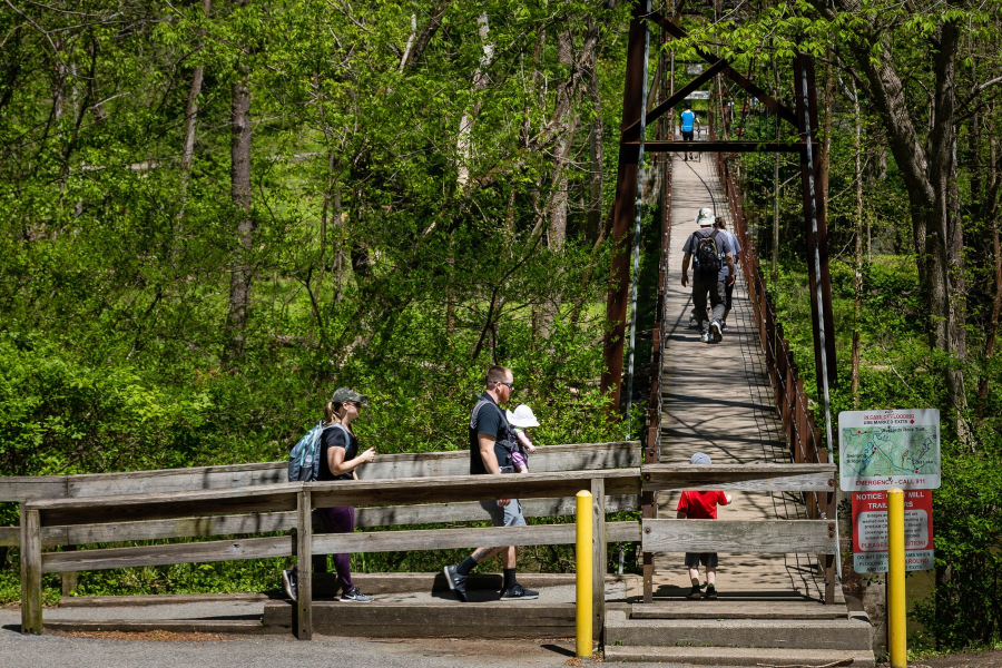 Hikers cross a swinging bridge over a river. Trees line the riverbank.