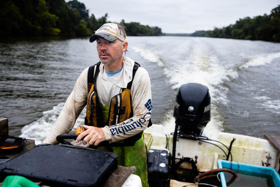 Balazik looks out ahead of himself while piloting his boat, creating white wake lines behind him on the river.