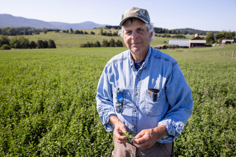 Mike wears a jean shirt, a hat, and has gray hair. He stands in front of his farm.