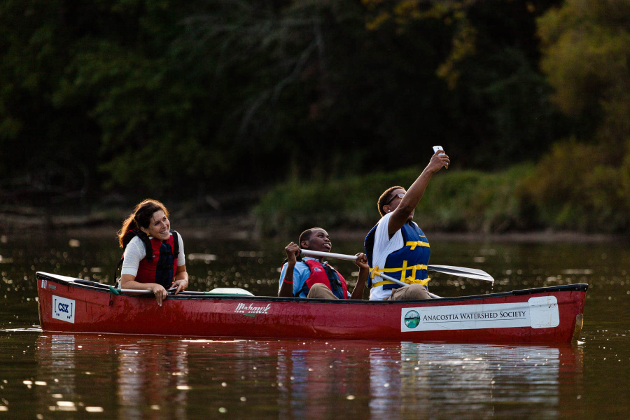 Two students and a guide pose for a selfie while paddling a canoe down a calm river at sunset.