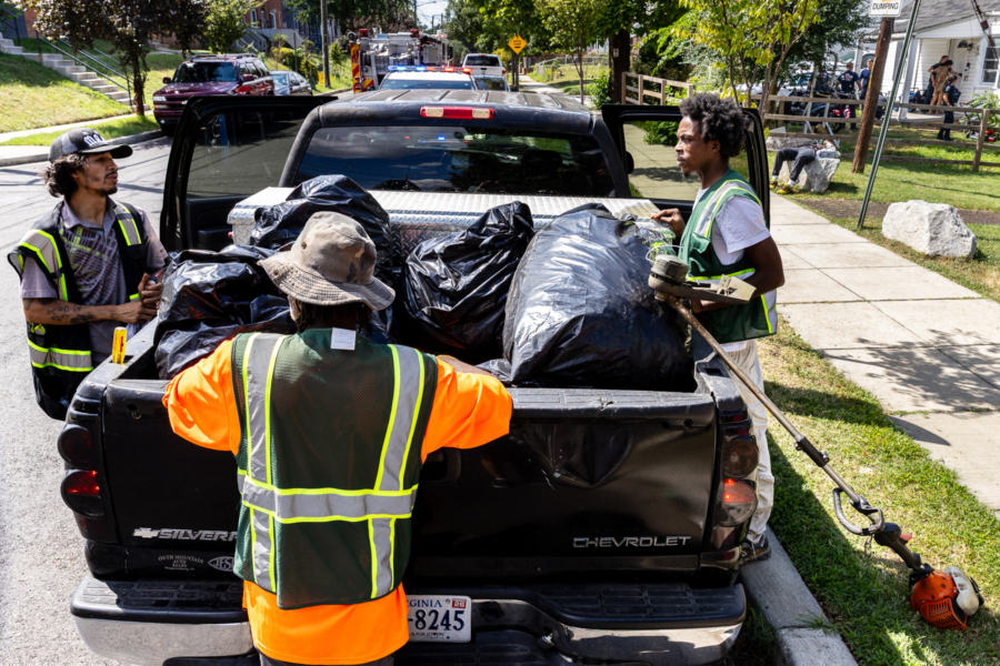 Three crew members rest on all sides of the bed of a pickup truck, police activity faintly visible in front of a house at a distance